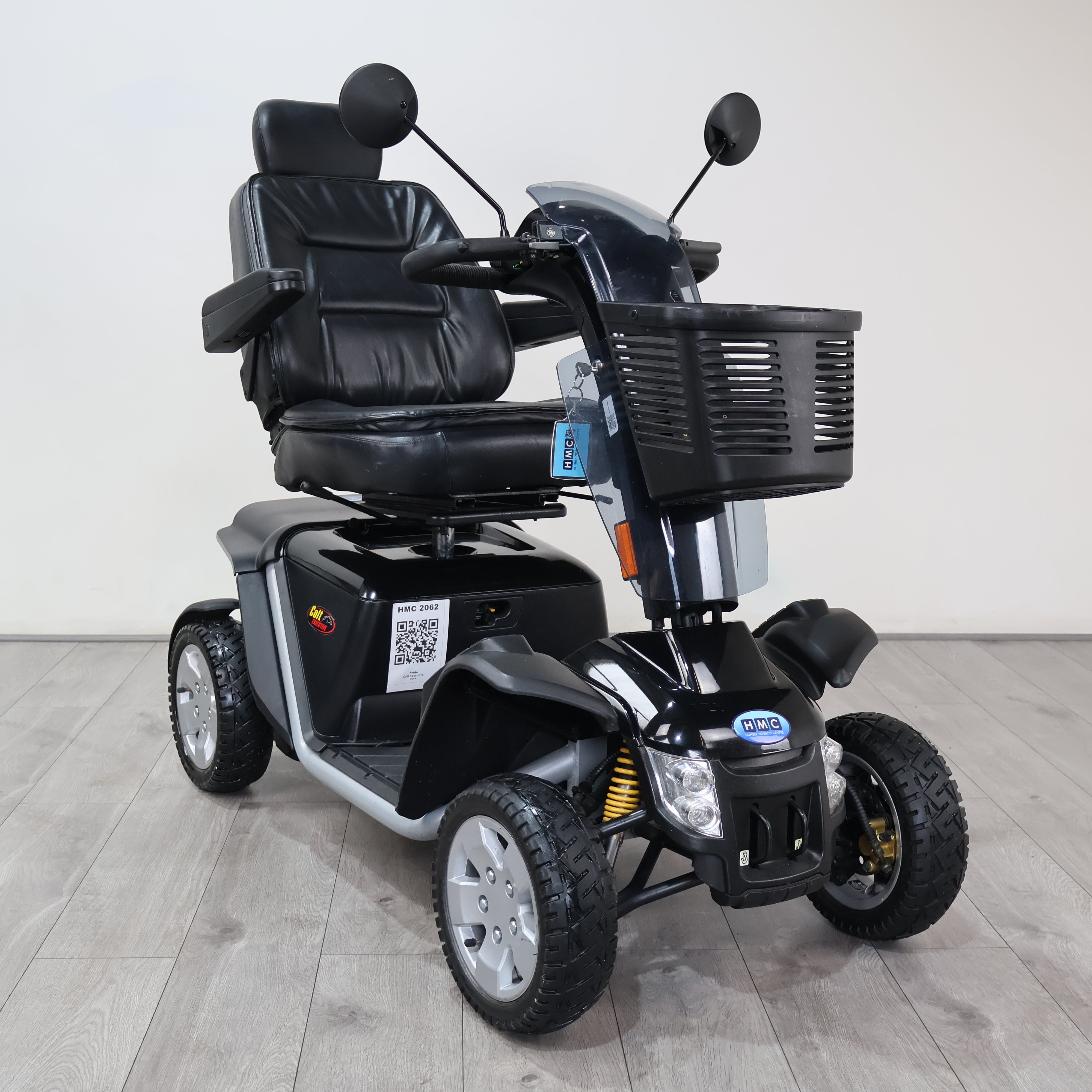 A black and gray three-wheel electric mobility scooter with a front basket, black leather-style seat, and large treaded tires, photographed indoors against a plain light wall and wood-look floor.