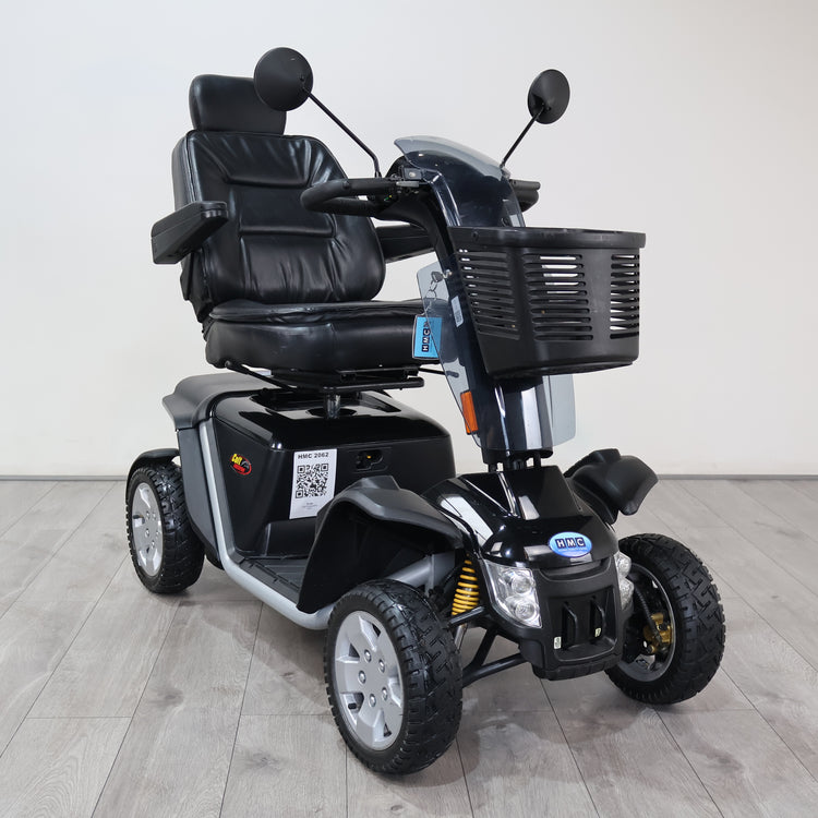 A black and gray three-wheel electric mobility scooter with a front basket, black leather-style seat, and large treaded tires, photographed indoors against a plain light wall and wood-look floor.