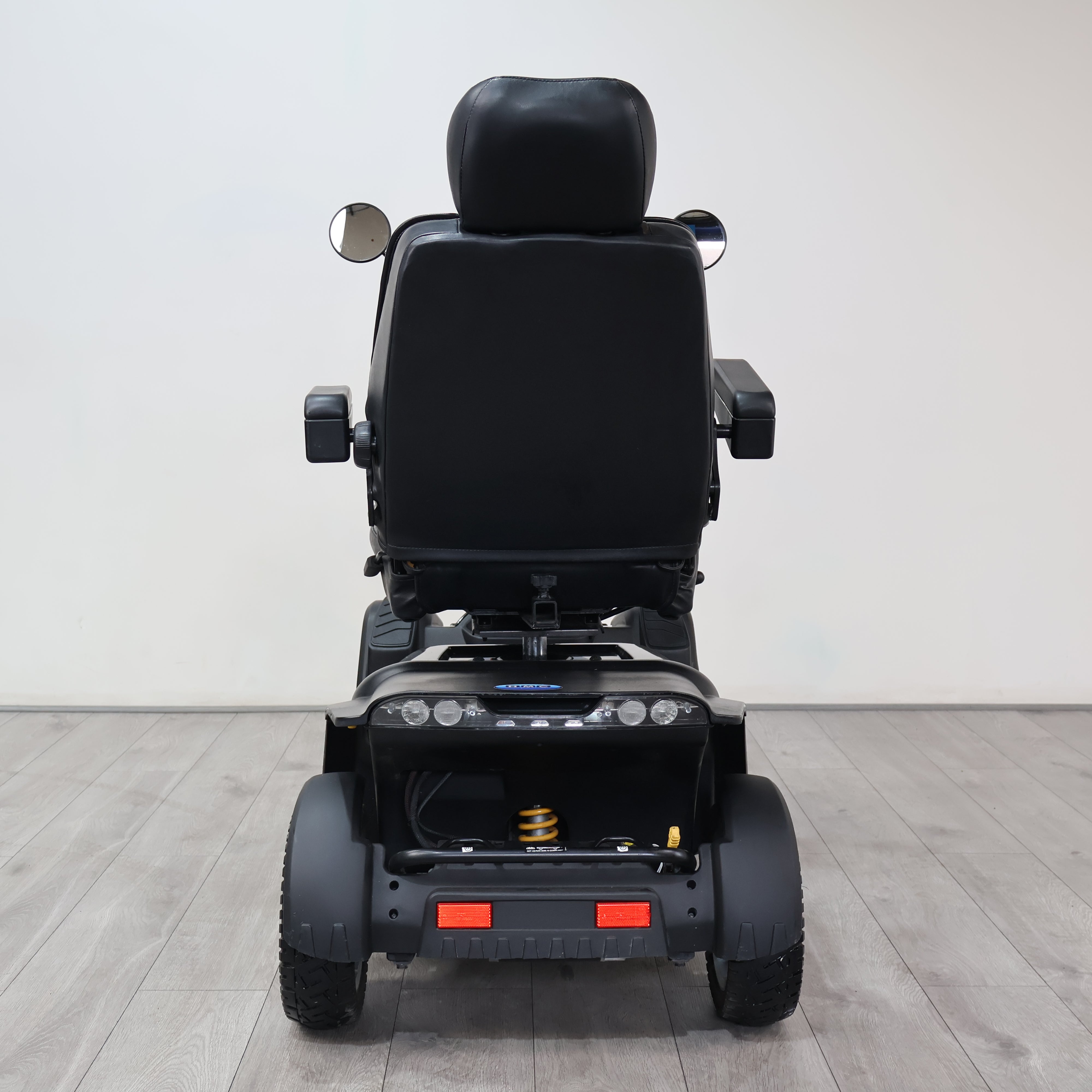 Rear view of a black power wheelchair with a padded seat, side mirrors, and rear wheels, photographed in a clean indoor studio setting with a light gray wall and wood-look floor.