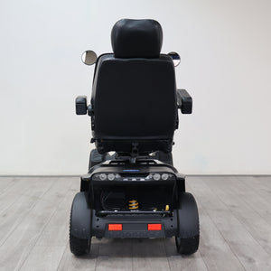 Rear view of a black power wheelchair with a padded seat, side mirrors, and rear wheels, photographed in a clean indoor studio setting with a light gray wall and wood-look floor.
