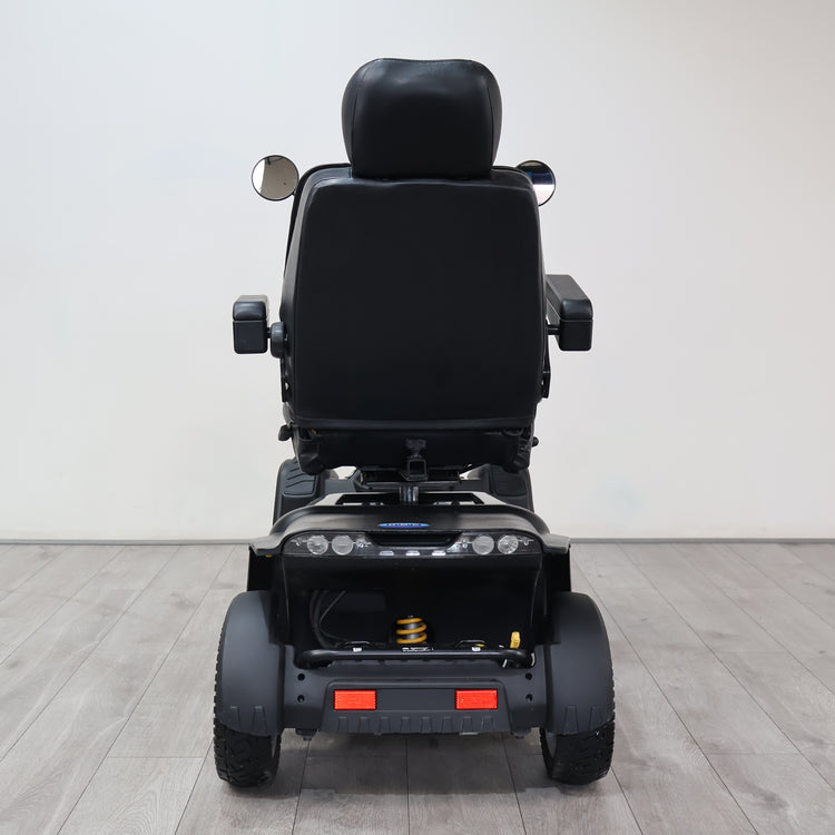 Rear view of a black power wheelchair with a padded seat, side mirrors, and rear wheels, photographed in a clean indoor studio setting with a light gray wall and wood-look floor.