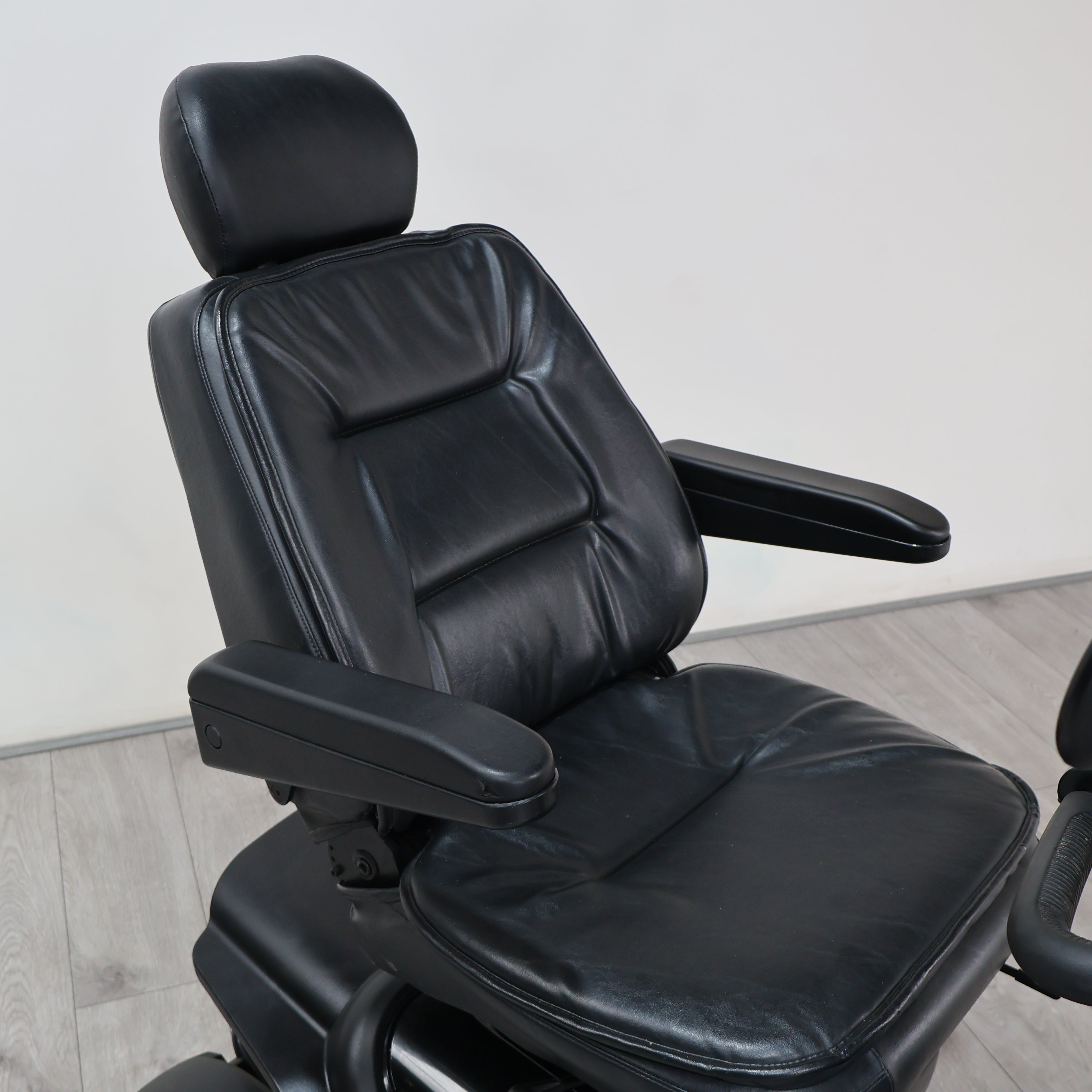 Black leather office chair with padded headrest, armrests, and a cushioned seat, photographed indoors against a plain light wall and wood floor.