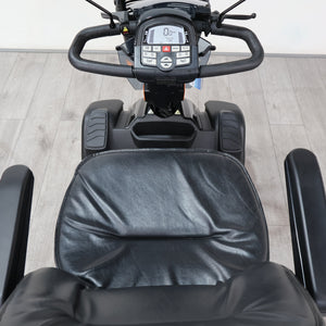 Black electric mobility scooter with a padded seat and digital control console, photographed from above on a light wood floor.