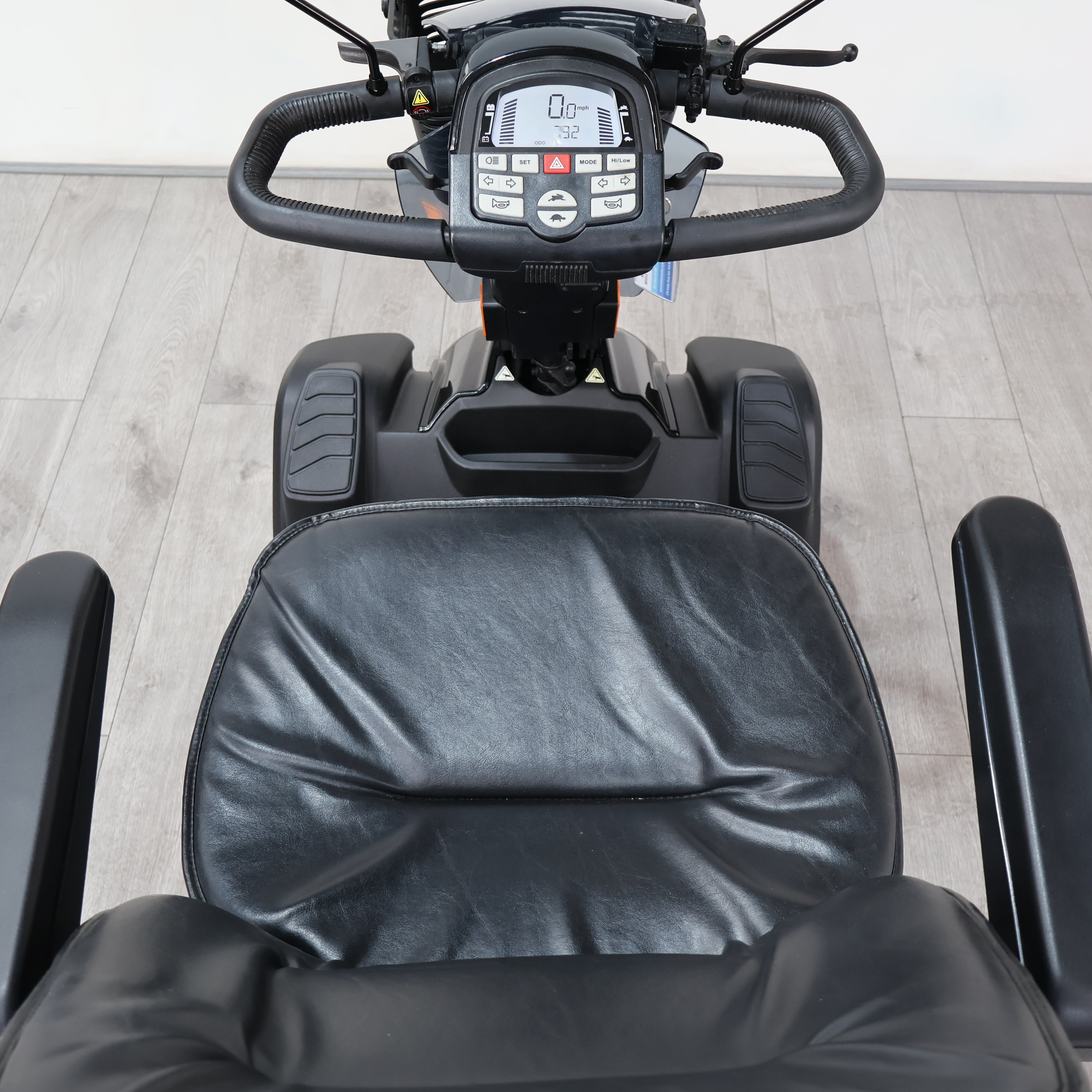 Black electric mobility scooter with a padded seat and digital control console, photographed from above on a light wood floor.