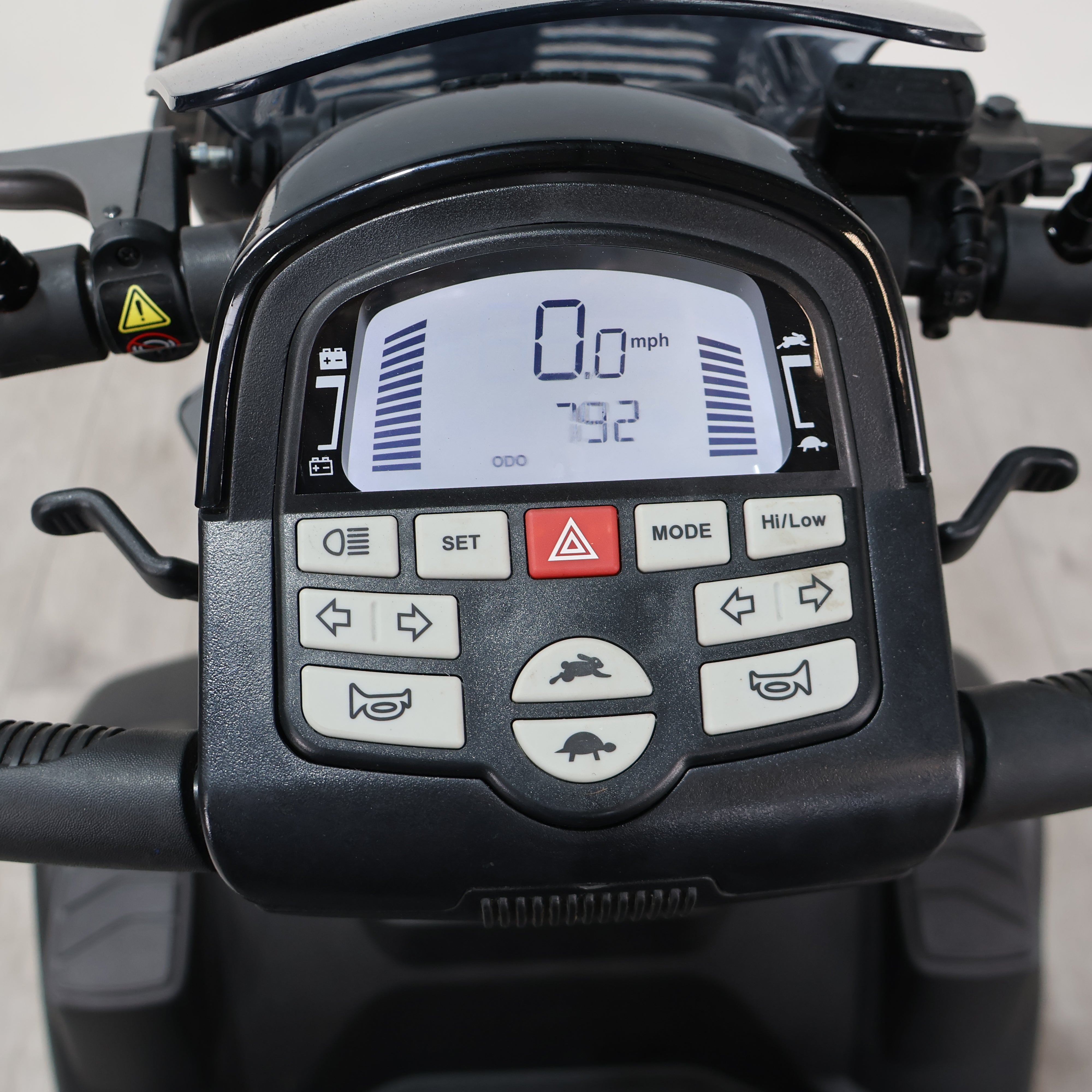 Close-up of a black electric scooter or moped handlebar control panel with a digital display and multiple buttons, mounted on the handlebars.