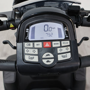 Close-up of a black electric scooter or moped handlebar control panel with a digital display and multiple buttons, mounted on the handlebars.