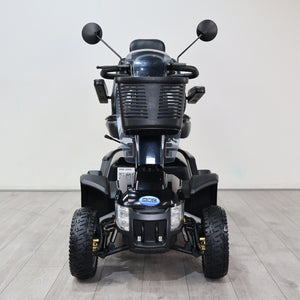 A black electric mobility quad bike with large off-road tires, a front storage basket, and side-view mirrors, photographed from the front in a clean indoor studio setting with a light gray wall and wood-look floor.
