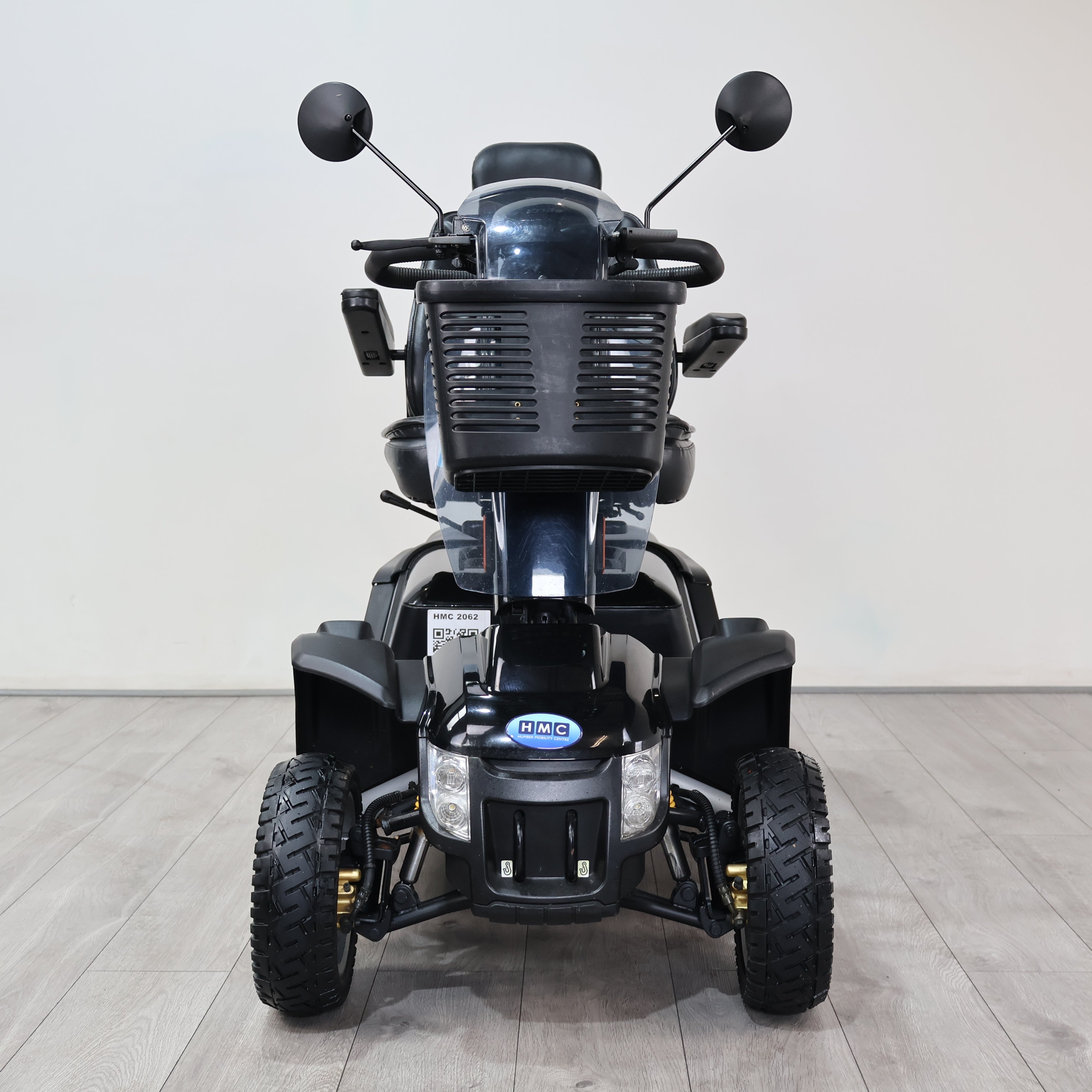 A black electric mobility quad bike with large off-road tires, a front storage basket, and side-view mirrors, photographed from the front in a clean indoor studio setting with a light gray wall and wood-look floor.