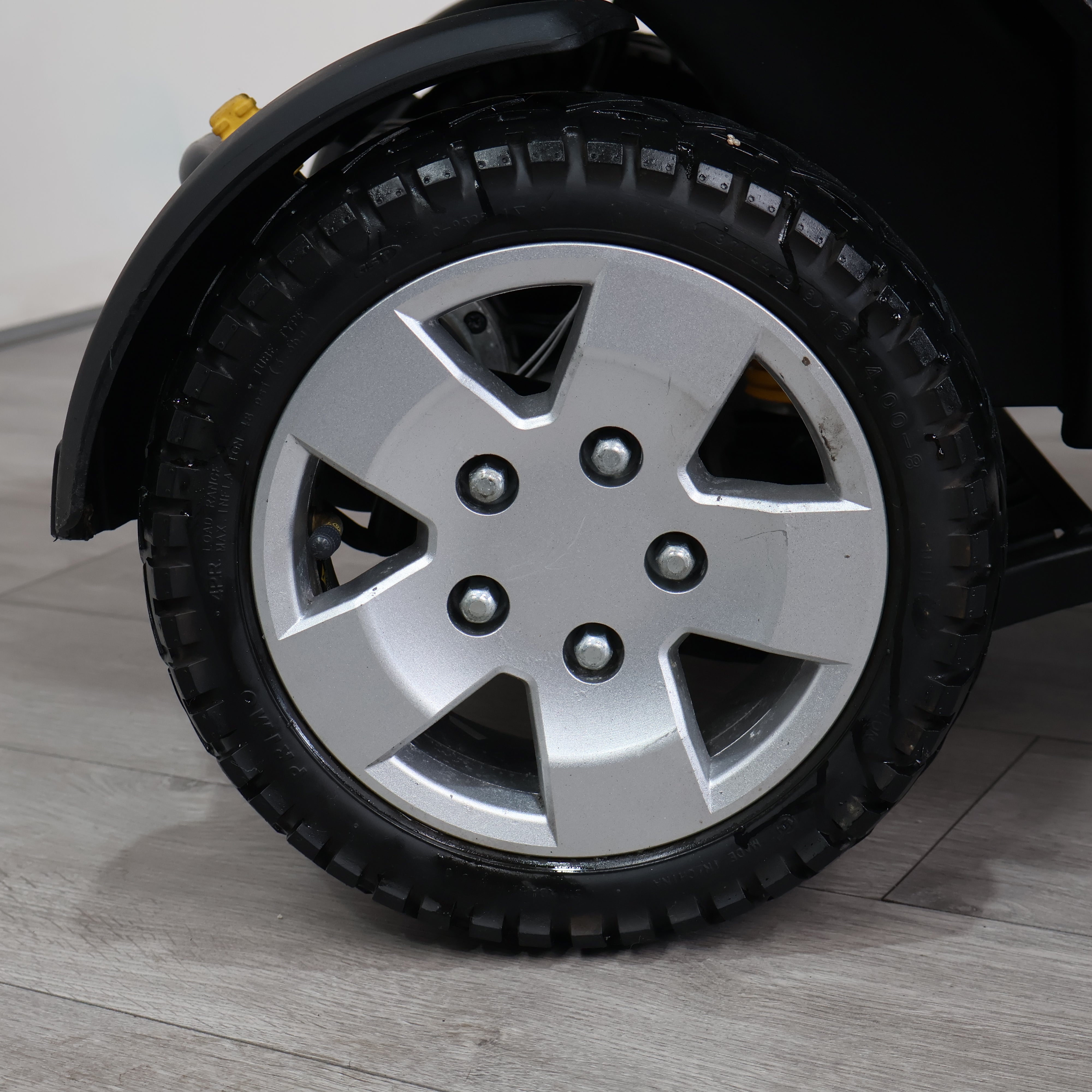 Close-up of a black tire with a silver five-spoke alloy wheel on a vehicle, photographed on a light gray floor against a white wall.