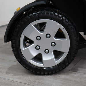 Close-up of a black tire with a silver five-spoke alloy wheel on a vehicle, photographed on a light gray floor against a white wall.