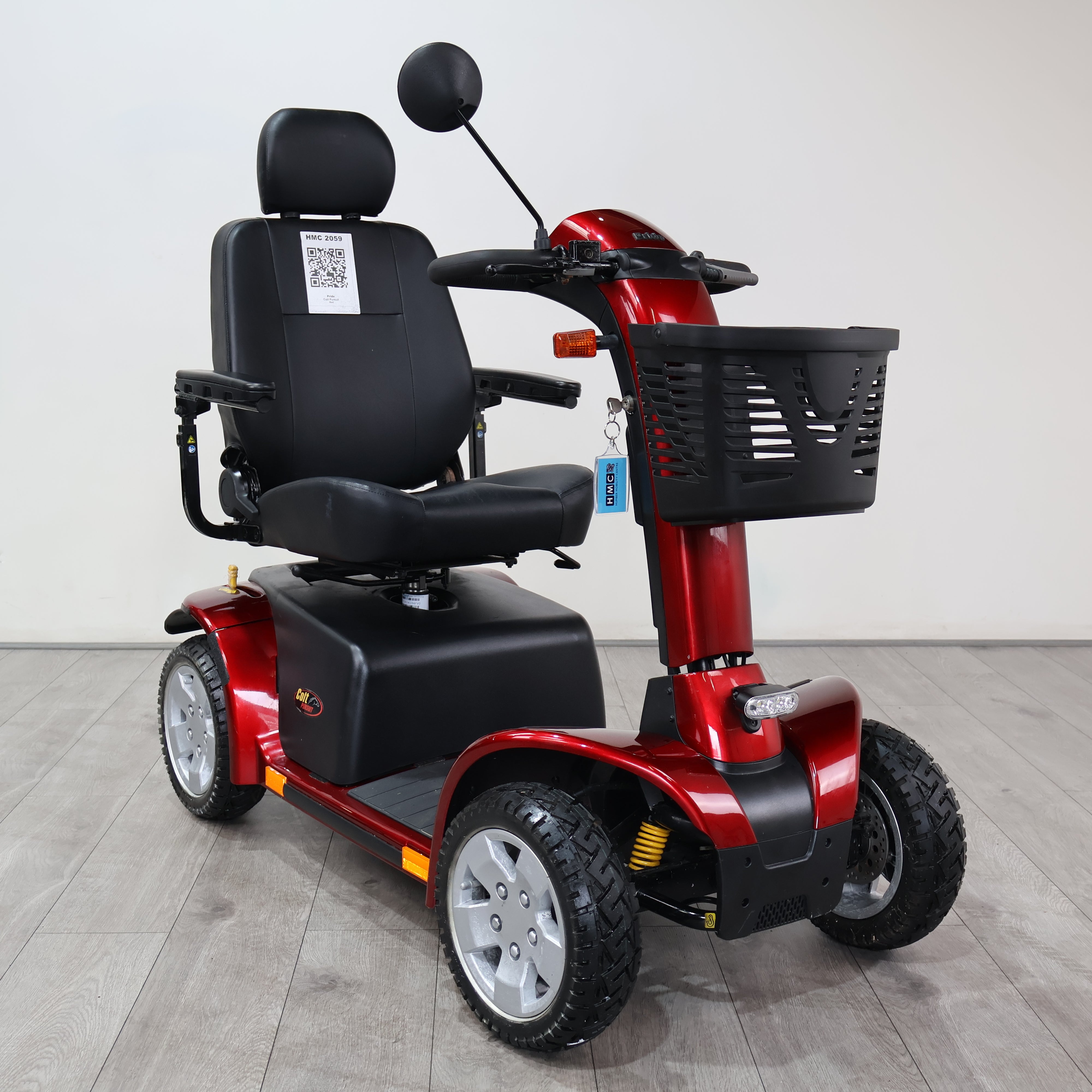A red and black four-wheel electric mobility scooter with a black seat, rearview mirror, and front basket, photographed indoors against a plain light gray wall and gray floor.