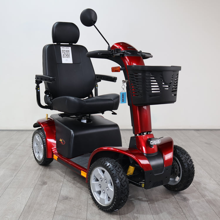 A red and black four-wheel electric mobility scooter with a black seat, rearview mirror, and front basket, photographed indoors against a plain light gray wall and gray floor.