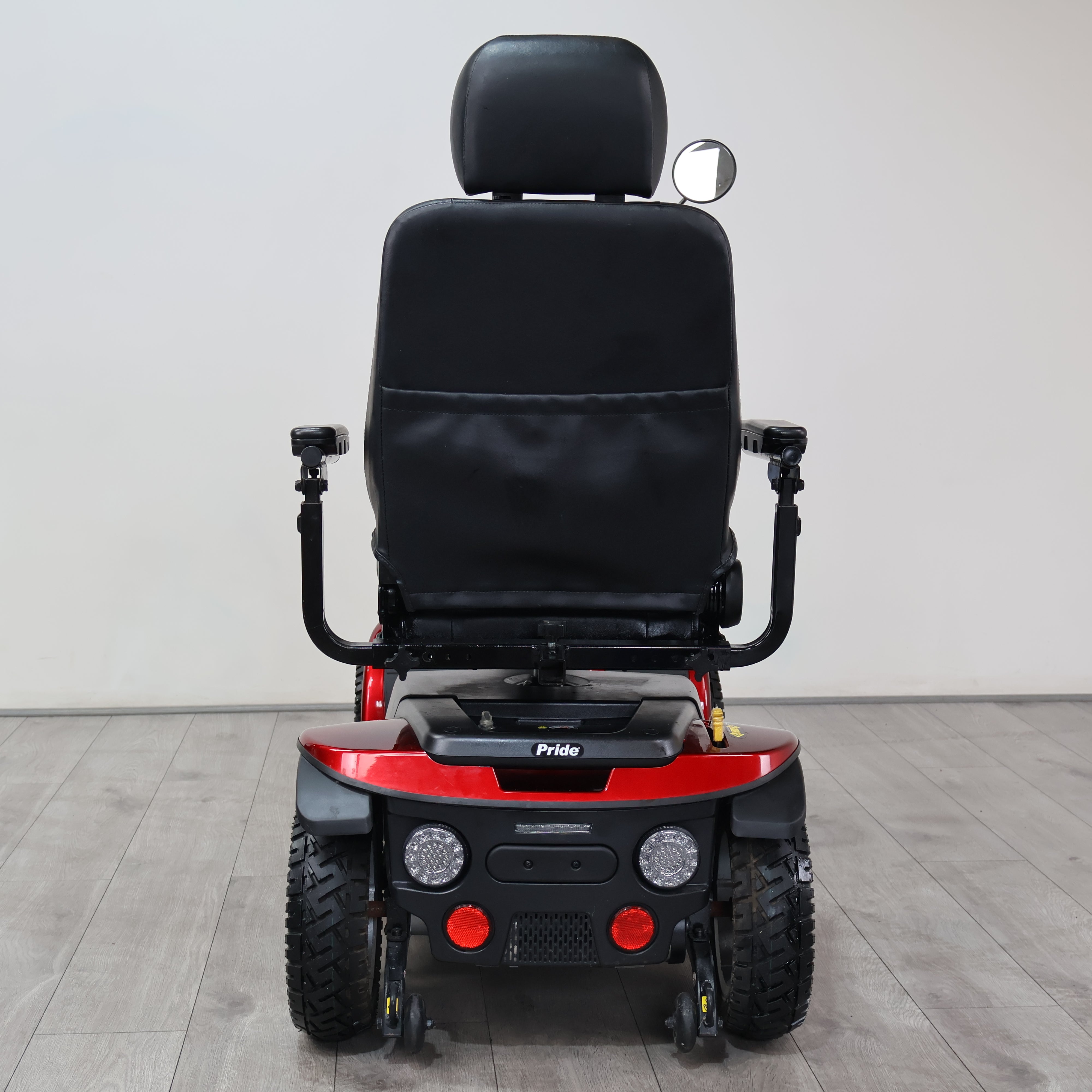 Rear view of a Pride electric power wheelchair with a black padded seat, black armrests, and a red body shell, photographed indoors against a light gray wall and gray floor.