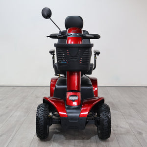 A red and black electric mobility scooter photographed from the rear, positioned on a light gray wood-look floor against a plain light gray wall.