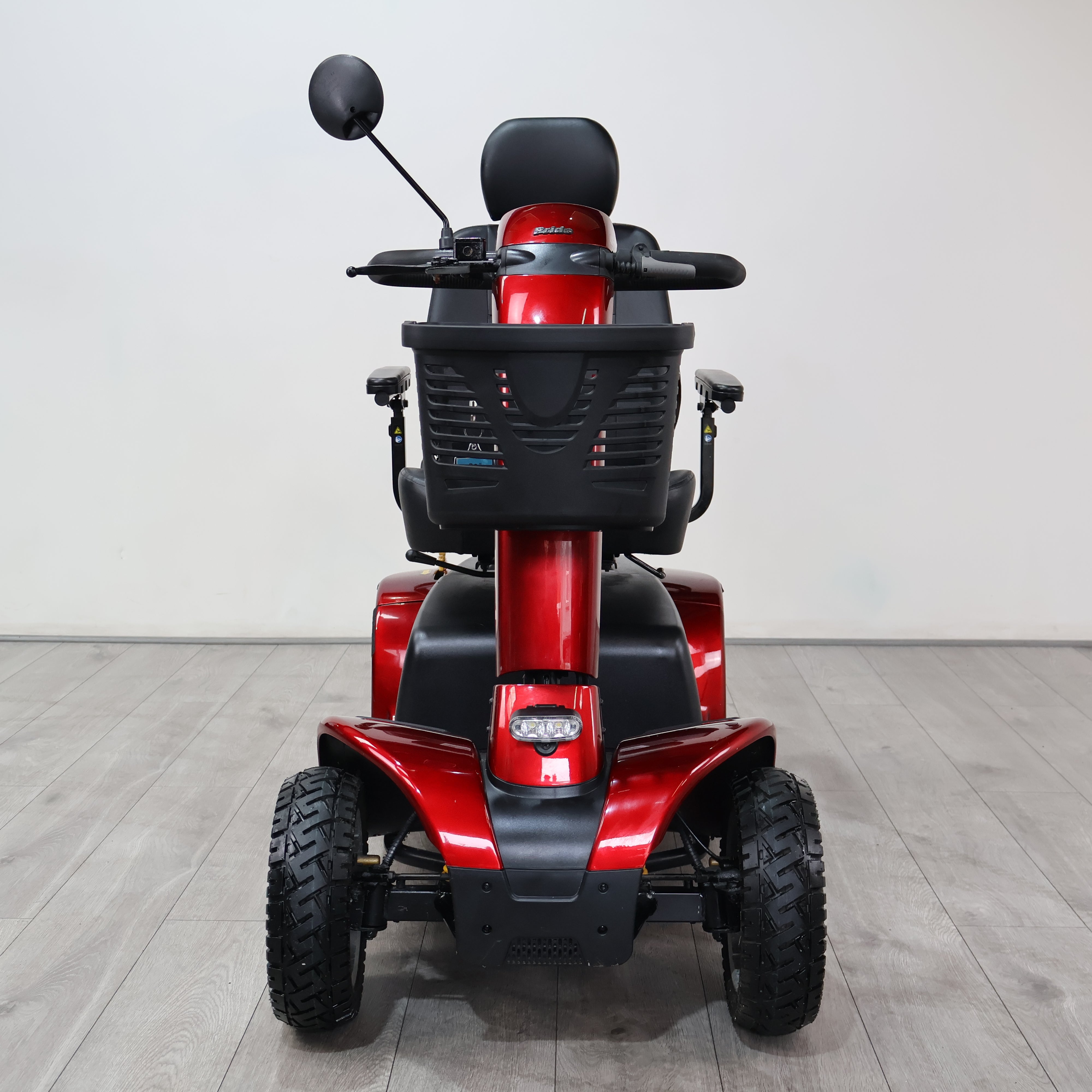 A red and black electric mobility scooter photographed from the rear, positioned on a light gray wood-look floor against a plain light gray wall.
