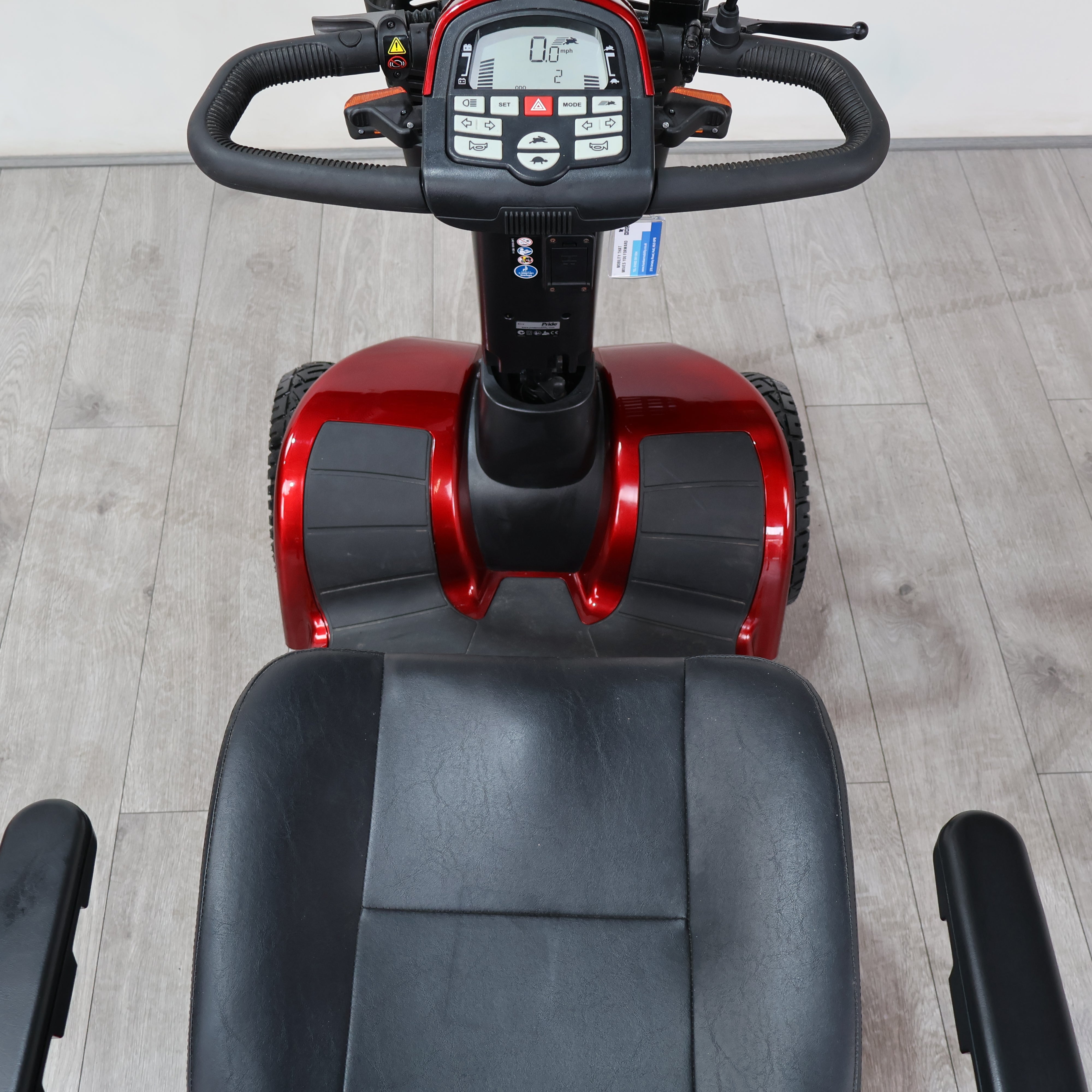An electric mobility scooter with a red and black body, featuring a black seat, handlebars, and a digital control console, photographed on a light gray wood-look floor.