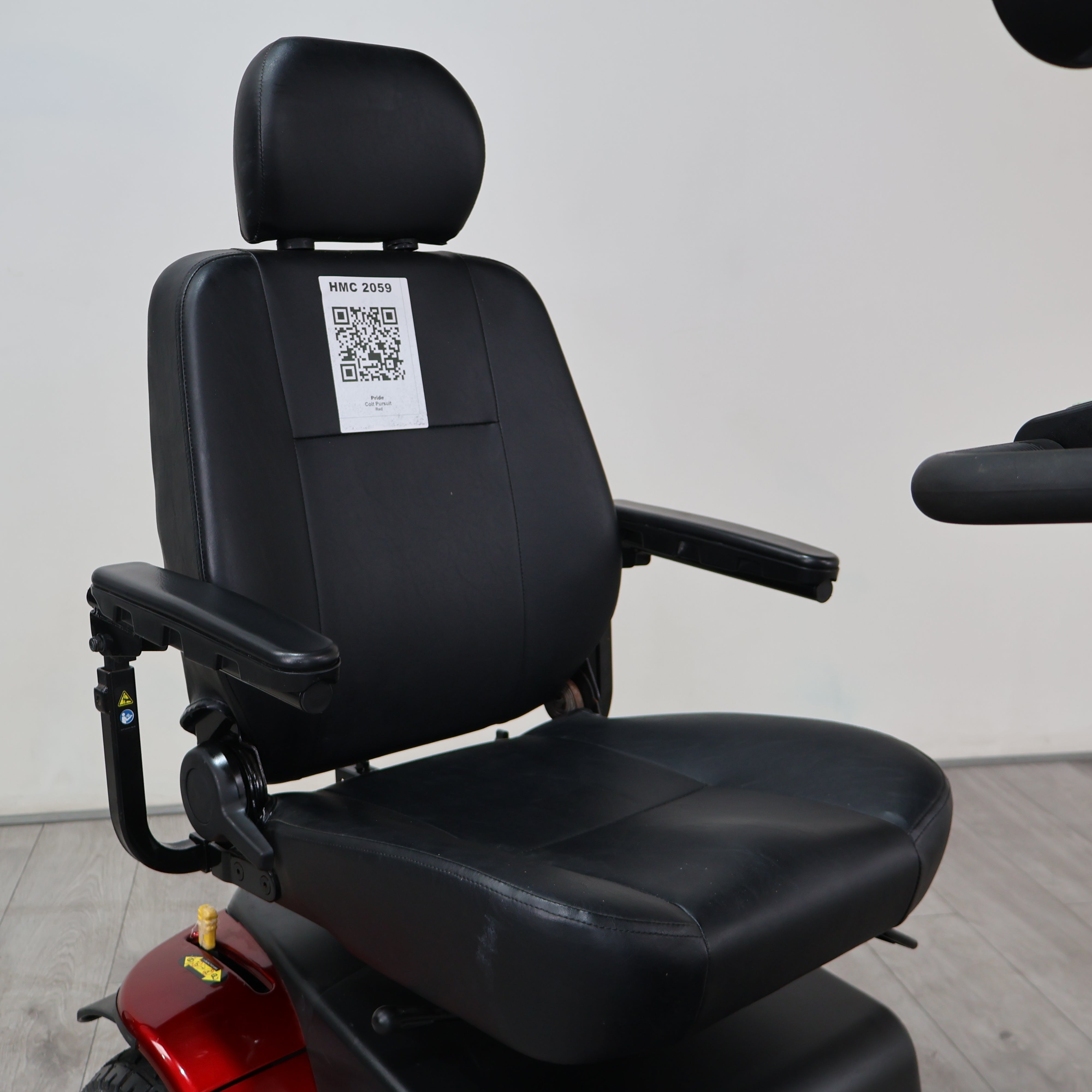 Black motorized scooter seat with headrest and armrests, photographed indoors against a plain light wall. A white QR code sticker is attached to the backrest, and part of a red vehicle body is visible underneath.