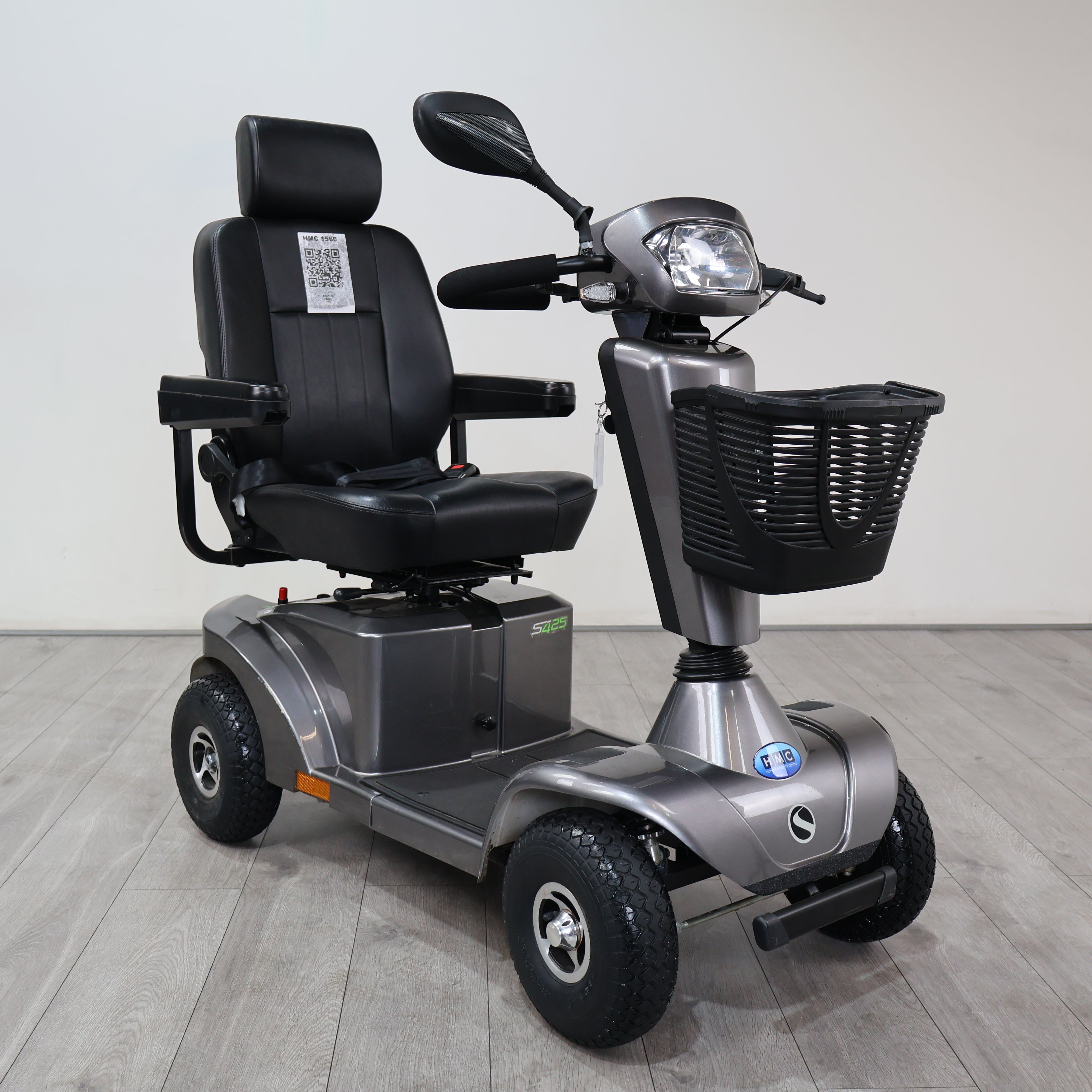 A silver and black electric four-wheel mobility scooter with a high-back black seat, front basket, and headlight, photographed indoors on a light gray floor against a plain light gray wall.