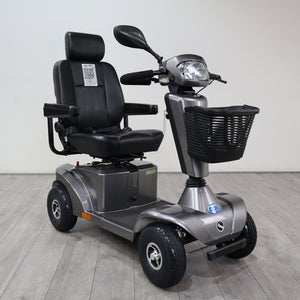 A silver and black electric four-wheel mobility scooter with a high-back black seat, front basket, and headlight, photographed indoors on a light gray floor against a plain light gray wall.