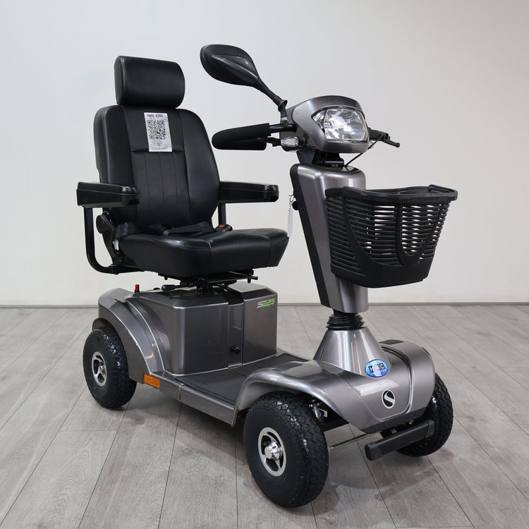 A silver and black electric four-wheel mobility scooter with a high-back black seat, front basket, and headlight, photographed indoors on a light gray floor against a plain light gray wall.