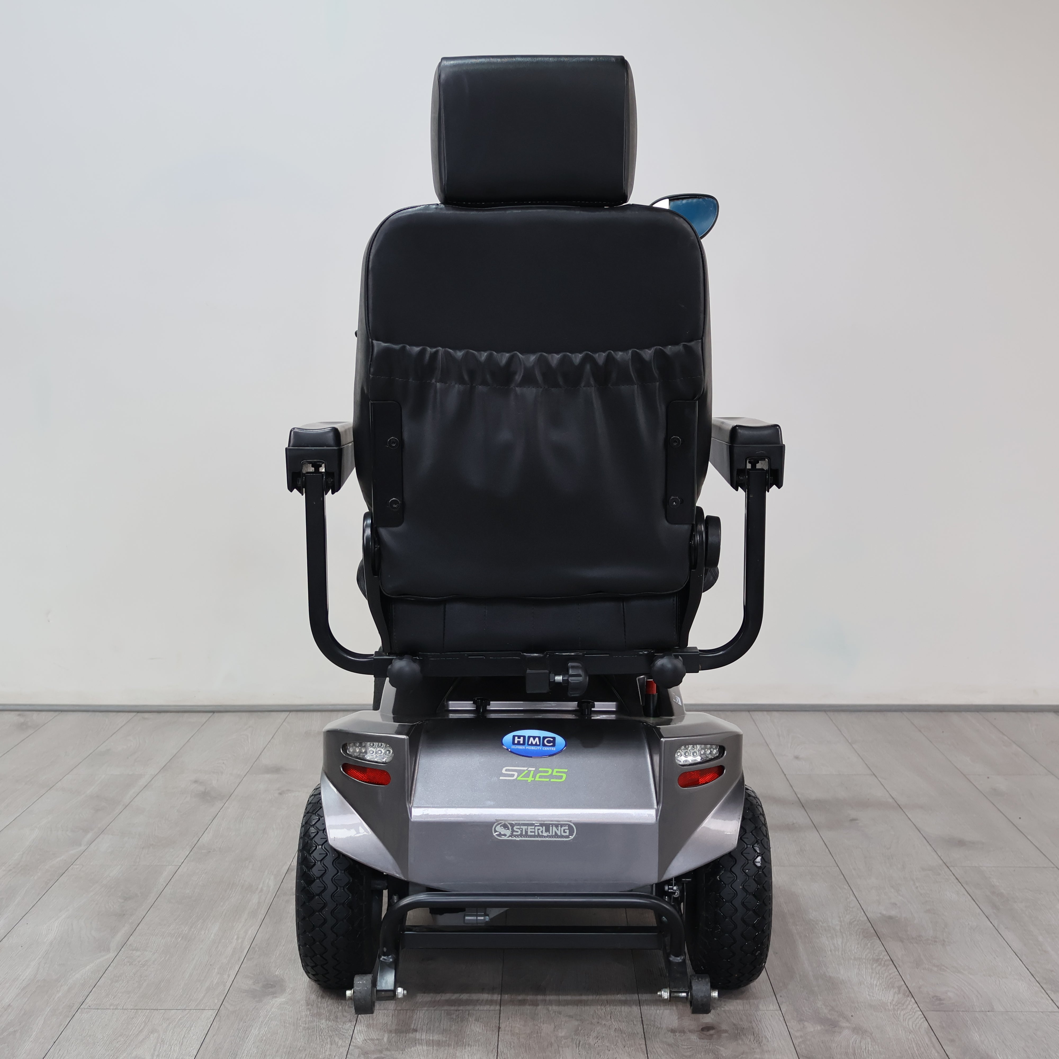 A rear view of a Power Sits electric mobility chair with a black padded seat and backrest, mounted on a silver motorized base with black wheels, set against a plain light gray wall and wood-look floor.