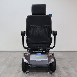 A rear view of a Power Sits electric mobility chair with a black padded seat and backrest, mounted on a silver motorized base with black wheels, set against a plain light gray wall and wood-look floor.