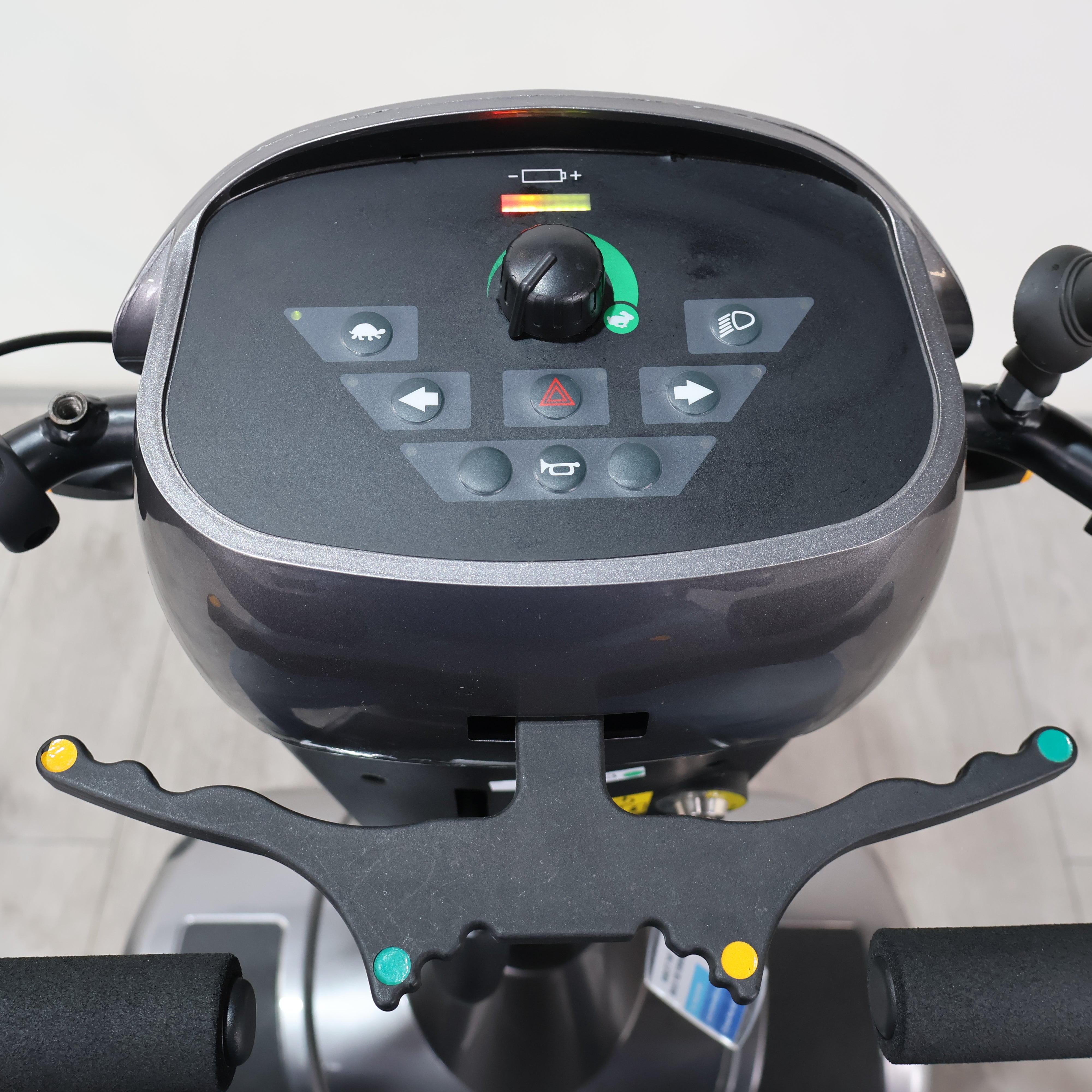 Close-up of a motorized scooter or exercise bike handlebar assembly, showing the black control panel with buttons, a central dial, and mounting arms with colored accents.