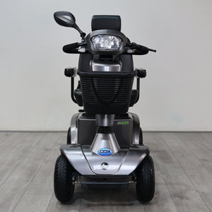 A silver and black electric mobility scooter with a headlight, rearview mirror, and three wheels, photographed from the front against a plain indoor wall and wood-look floor.