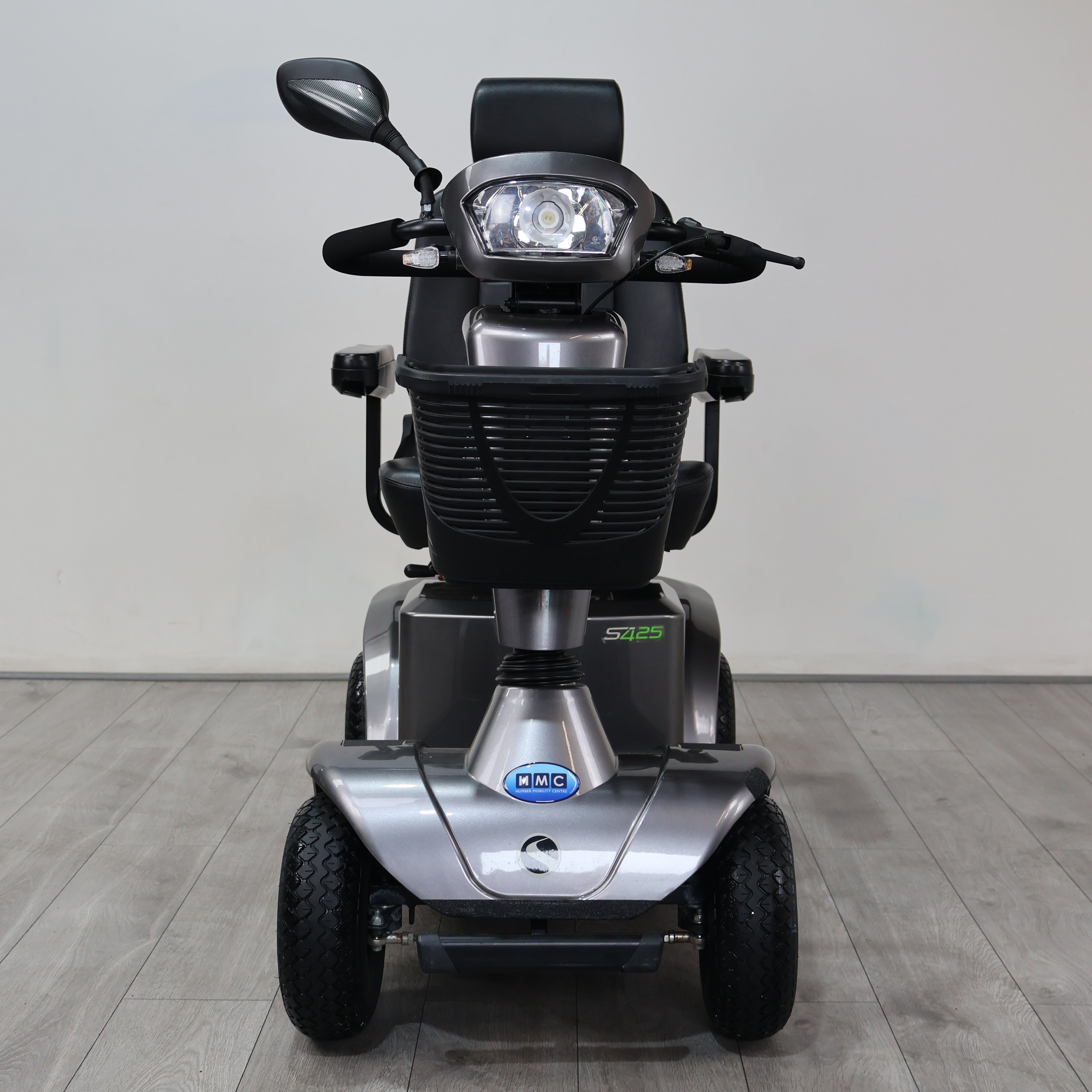 A silver and black electric mobility scooter with a headlight, rearview mirror, and three wheels, photographed from the front against a plain indoor wall and wood-look floor.
