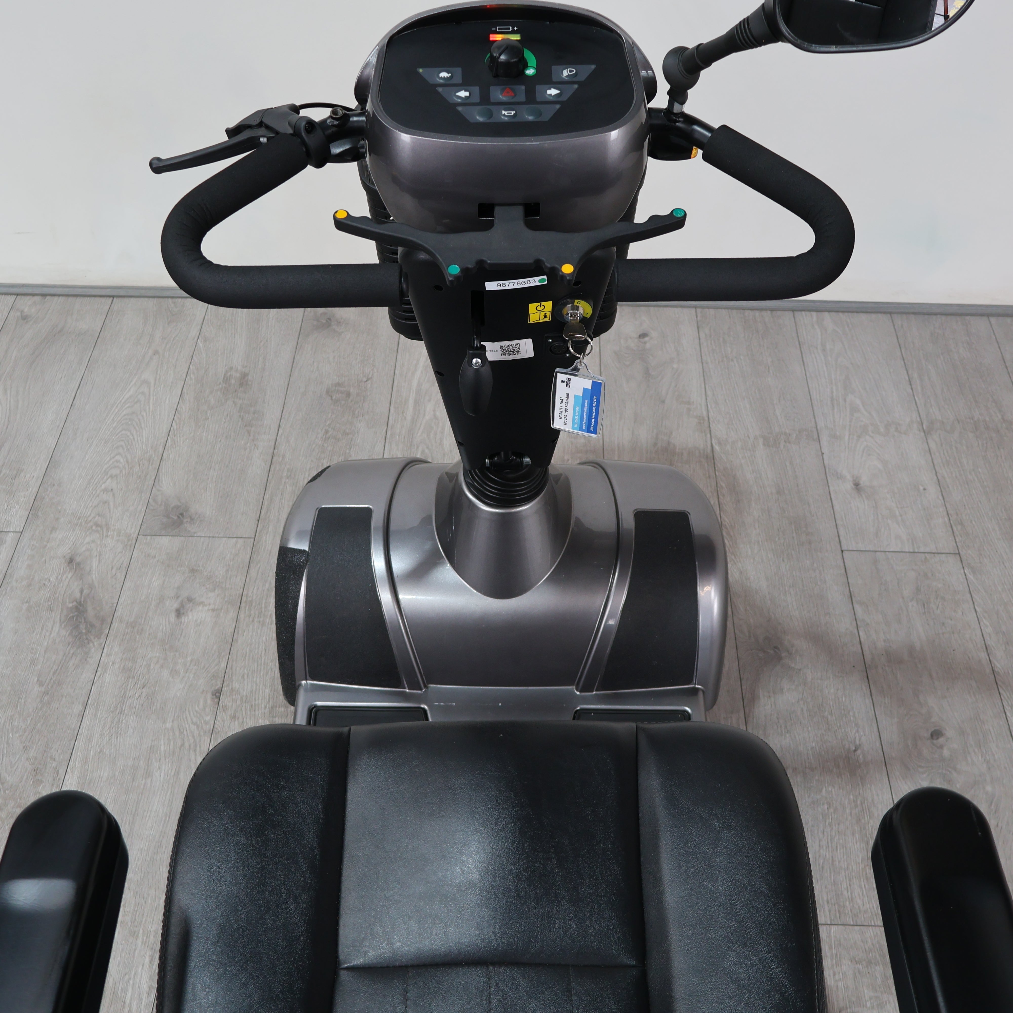 A personal exercise vehicle with a black seat, black handlebars, and a gray and black control console, photographed indoors on a light wood floor against a white wall.