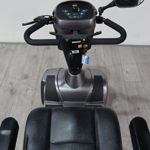 A personal exercise vehicle with a black seat, black handlebars, and a gray and black control console, photographed indoors on a light wood floor against a white wall.