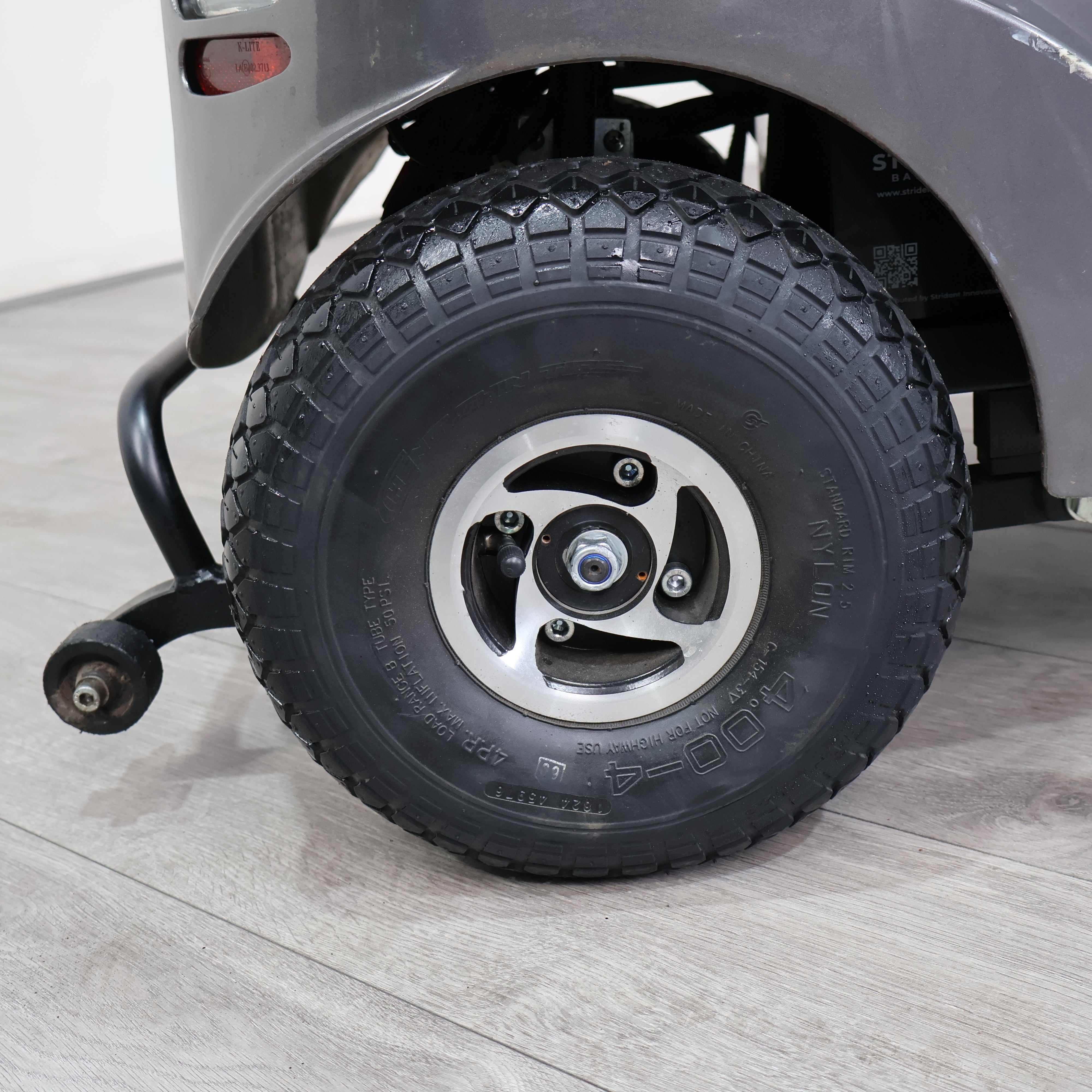 Close-up of a black all-terrain tire mounted on a silver five-spoke wheel, attached to a gray vehicle on a light gray wood-look floor.