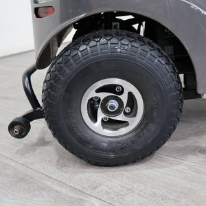 Close-up of a black all-terrain tire mounted on a silver five-spoke wheel, attached to a gray vehicle on a light gray wood-look floor.