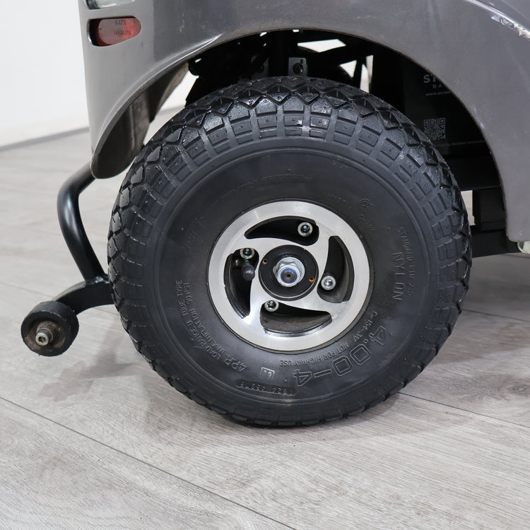 Close-up of a black all-terrain tire mounted on a silver five-spoke wheel, attached to a gray vehicle on a light gray wood-look floor.