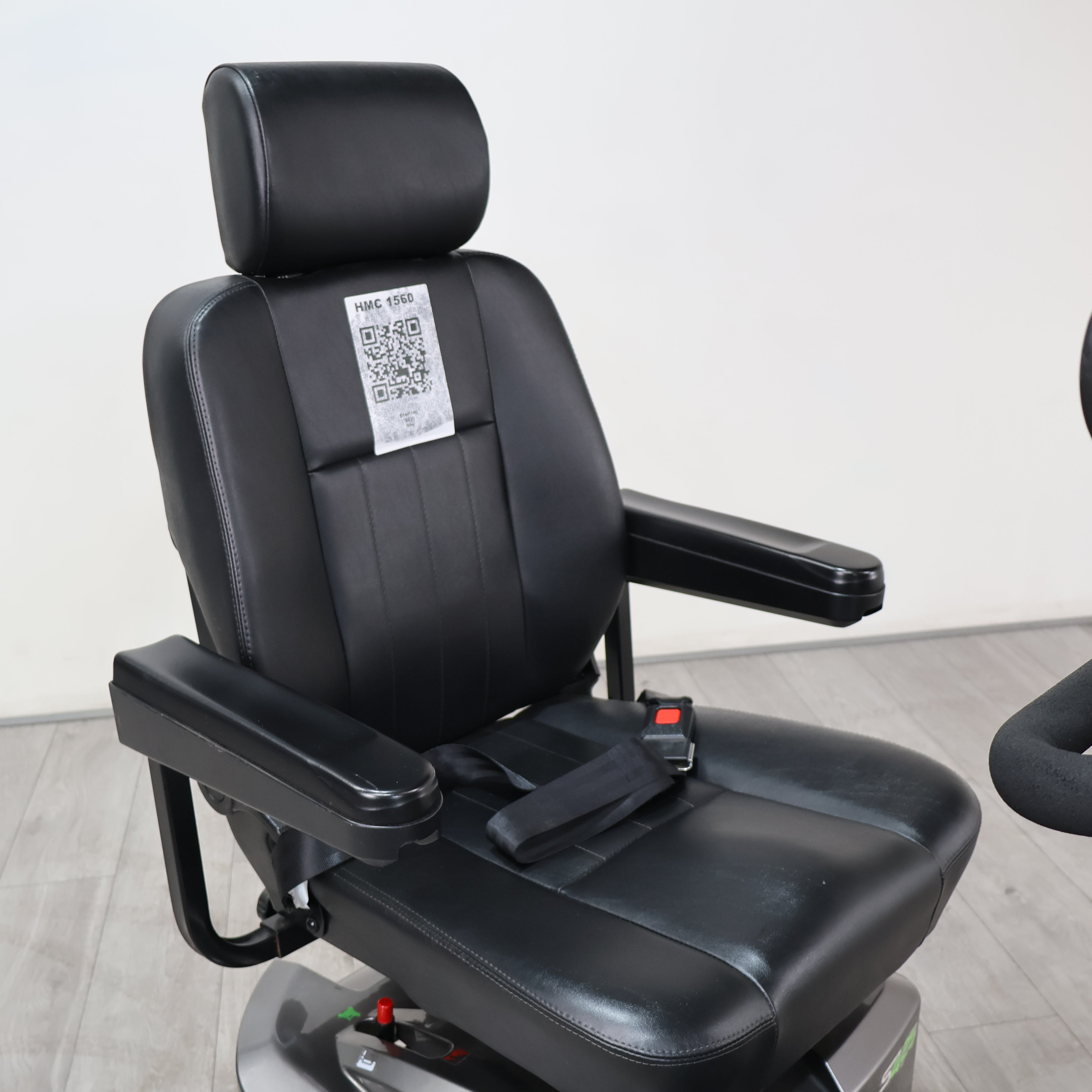 Black faux leather driver's seat with integrated armrests, a headrest, and a seat belt, shown in a showroom setting.