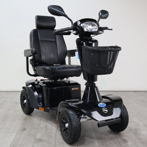 A black electric three-wheel mobility scooter with a padded seat, rear storage box, front basket, and headlight, photographed indoors on a light gray floor against a plain light wall.