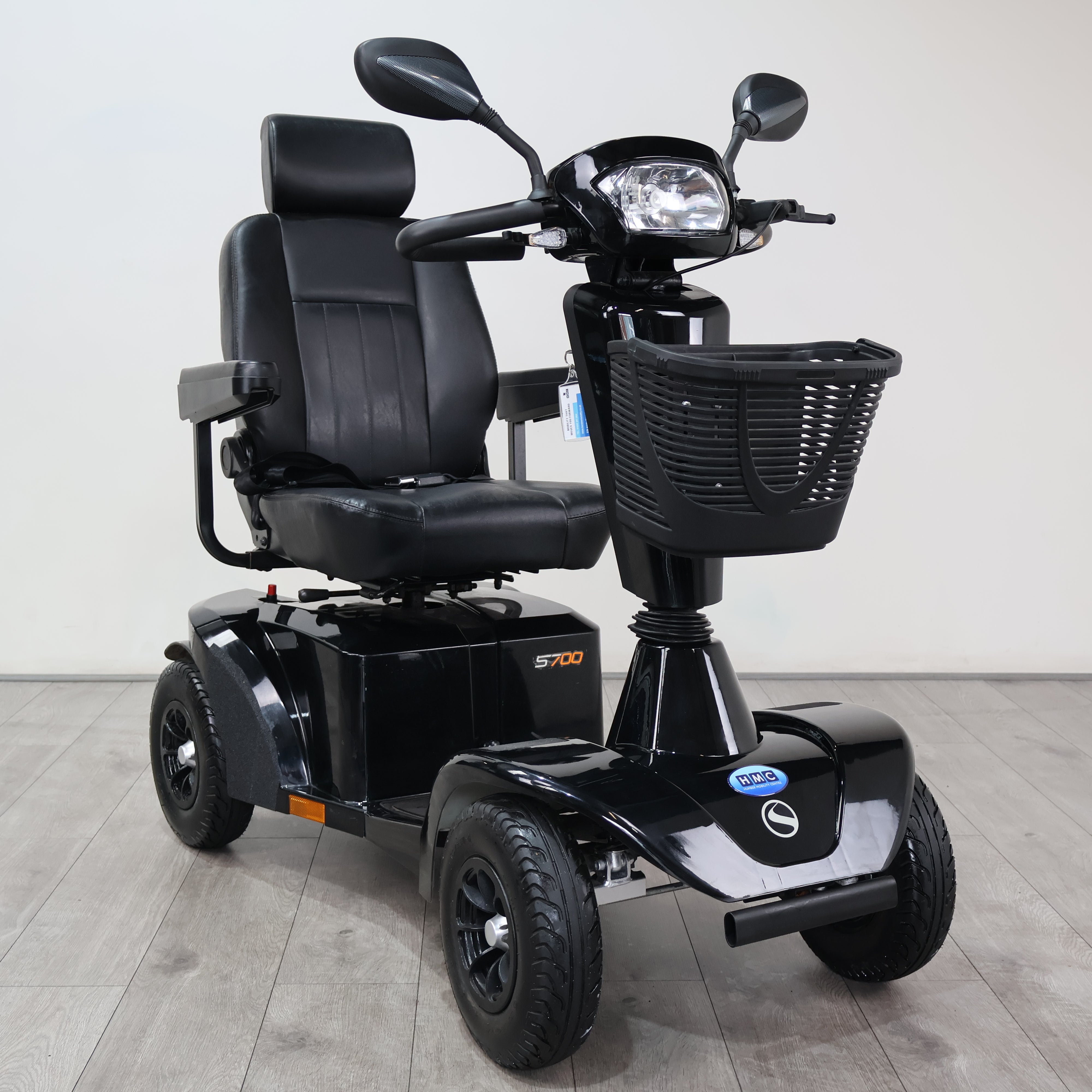 A black electric three-wheel mobility scooter with a padded seat, rear storage box, front basket, and headlight, photographed indoors on a light gray floor against a plain light wall.