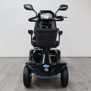 Black electric three-wheel mobility scooter with a front headlight, rearview mirrors, and a front storage basket, shown from the front on a light gray tiled floor against a white wall.