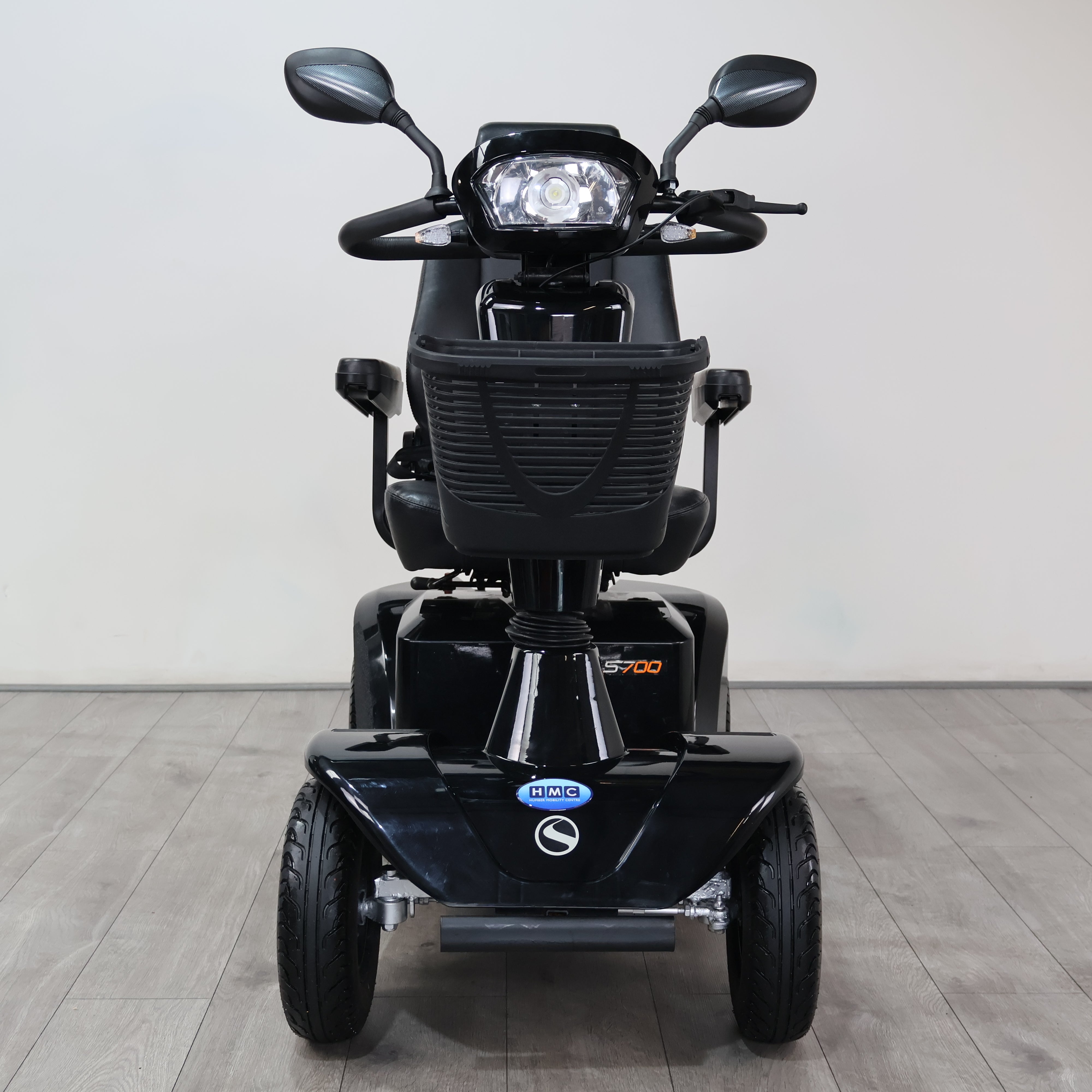 Black electric three-wheel mobility scooter with a front headlight, rearview mirrors, and a front storage basket, shown from the front on a light gray tiled floor against a white wall.