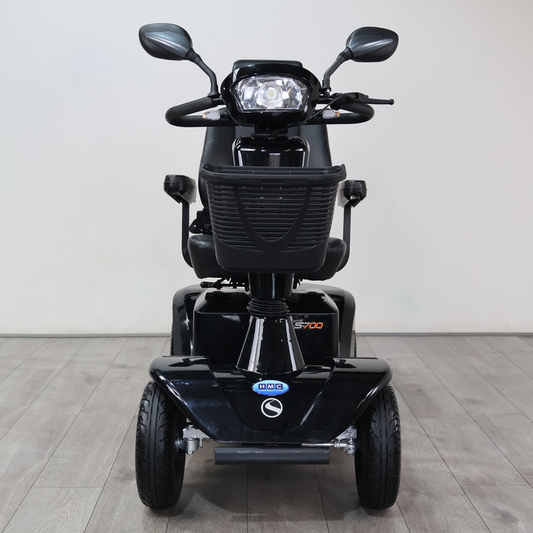 Black electric three-wheel mobility scooter with a front headlight, rearview mirrors, and a front storage basket, shown from the front on a light gray tiled floor against a white wall.