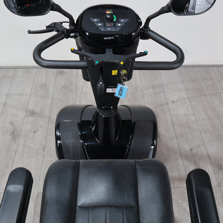 A black Segway-style personal transporter with a digital display screen, handlebars, and a black seat, photographed indoors on a light wood floor against a plain wall.