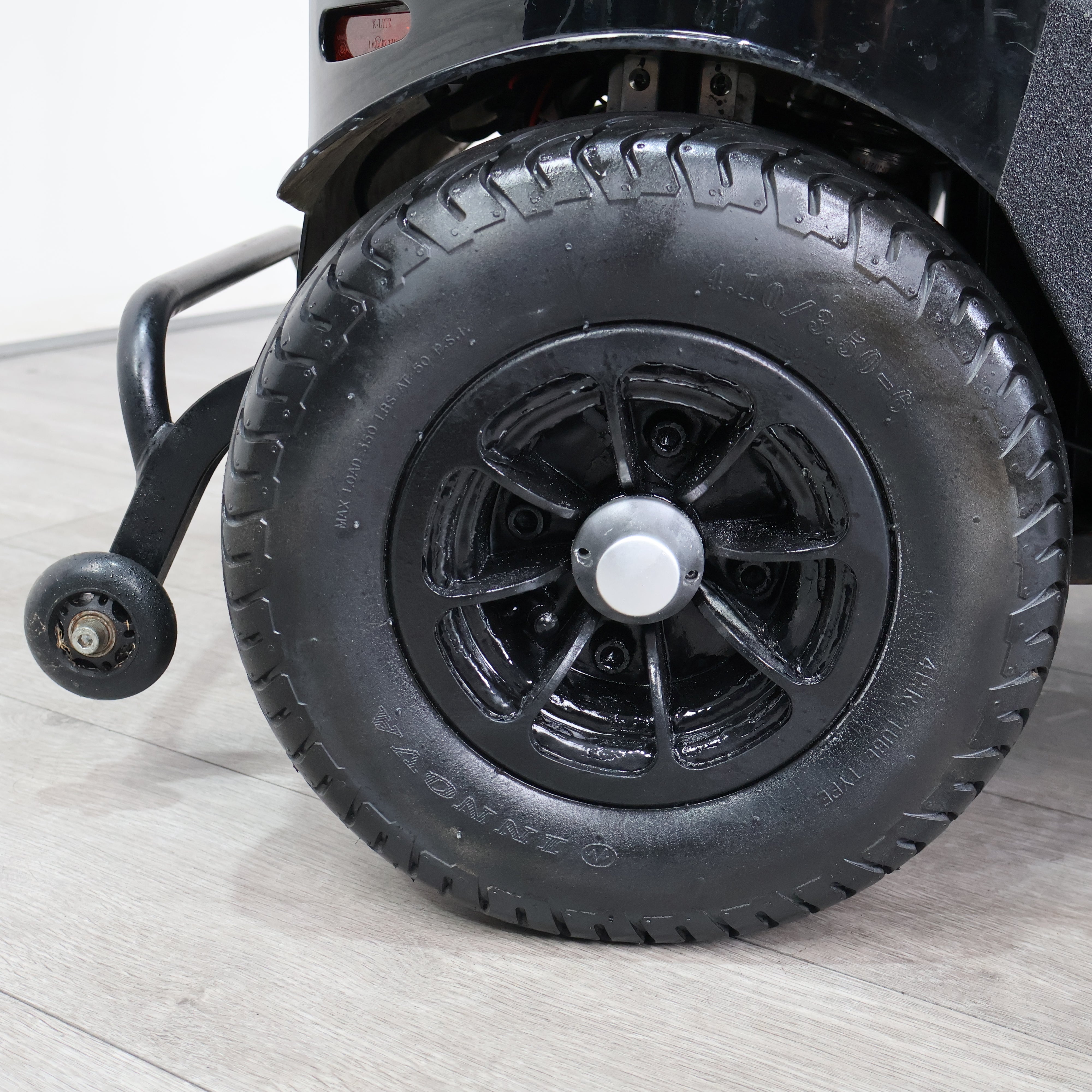 Close-up of a black rugged tire mounted on a black spoked wheel with a silver center cap, attached to a black vehicle on a light wood floor.