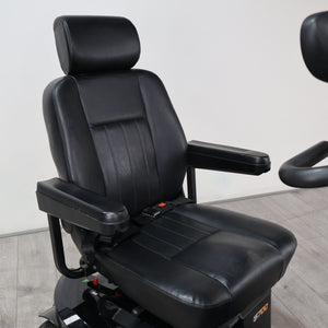 Black leather motorcycle or racing bucket seat with integrated armrests and a headrest, mounted on a black base with a visible seatbelt buckle area, photographed indoors against a plain light wall and gray floor.