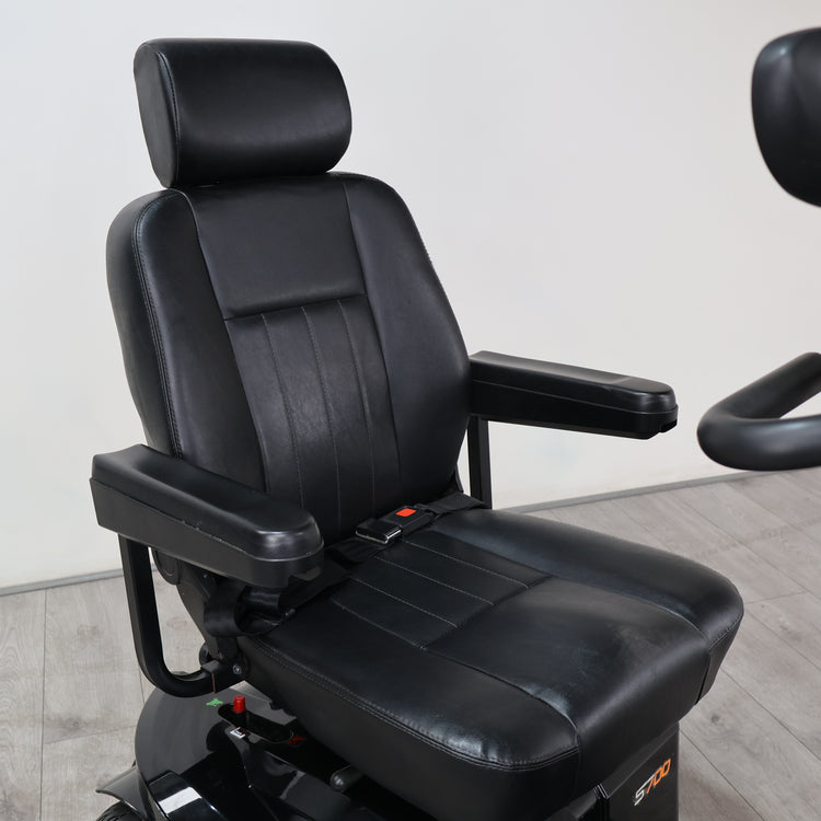 Black leather motorcycle or racing bucket seat with integrated armrests and a headrest, mounted on a black base with a visible seatbelt buckle area, photographed indoors against a plain light wall and gray floor.