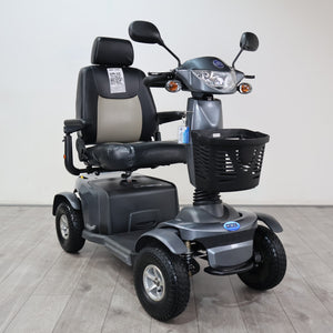 A gray and black four-wheel electric mobility scooter with a front basket, rear storage box, and a high-back seat, photographed indoors against a plain light wall and gray floor.