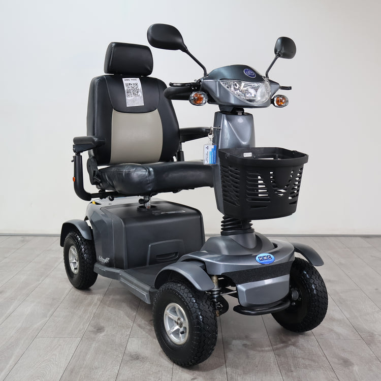 A gray and black four-wheel electric mobility scooter with a front basket, rear storage box, and a high-back seat, photographed indoors against a plain light wall and gray floor.
