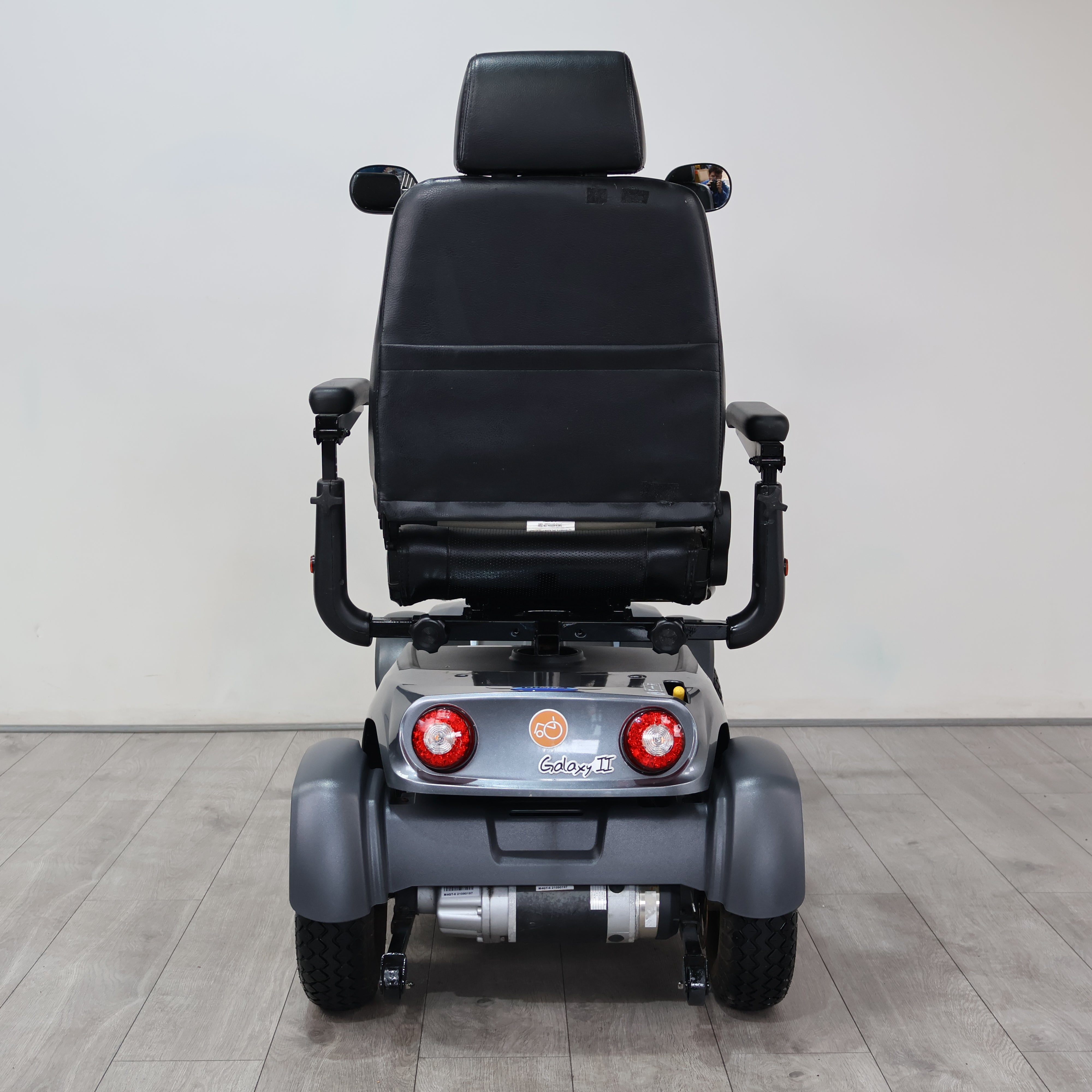A rear view of a motorized power wheelchair with a black padded seat and gray body, positioned on a light gray tiled floor against a plain light gray wall.