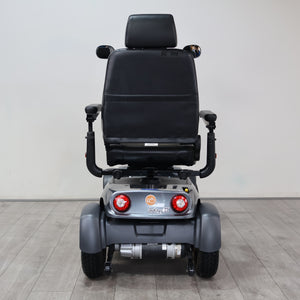 A rear view of a motorized power wheelchair with a black padded seat and gray body, positioned on a light gray tiled floor against a plain light gray wall.