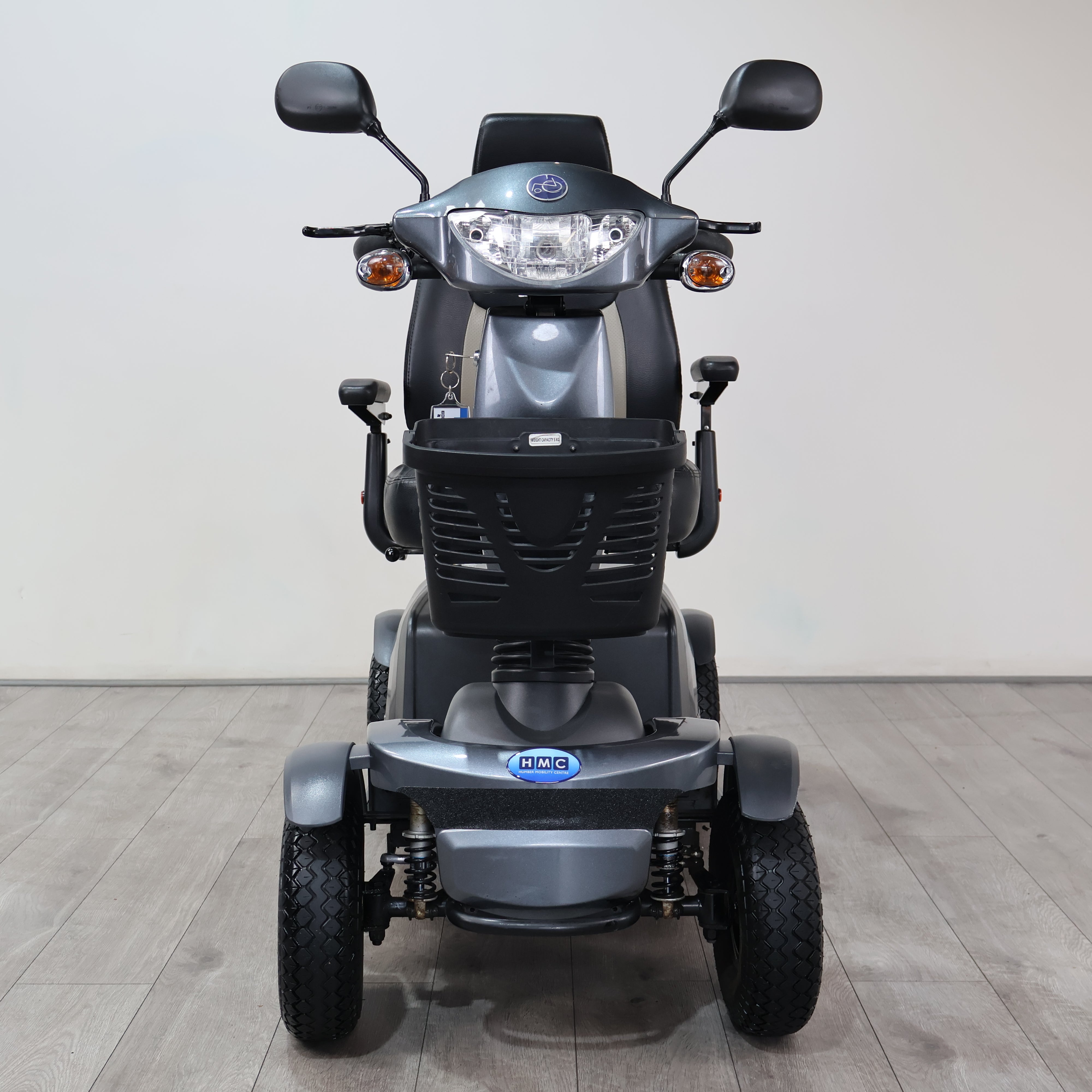 A gray and black IHQ electric three-wheel mobility scooter photographed from the front, set against a plain light gray wall and gray wood-look floor.