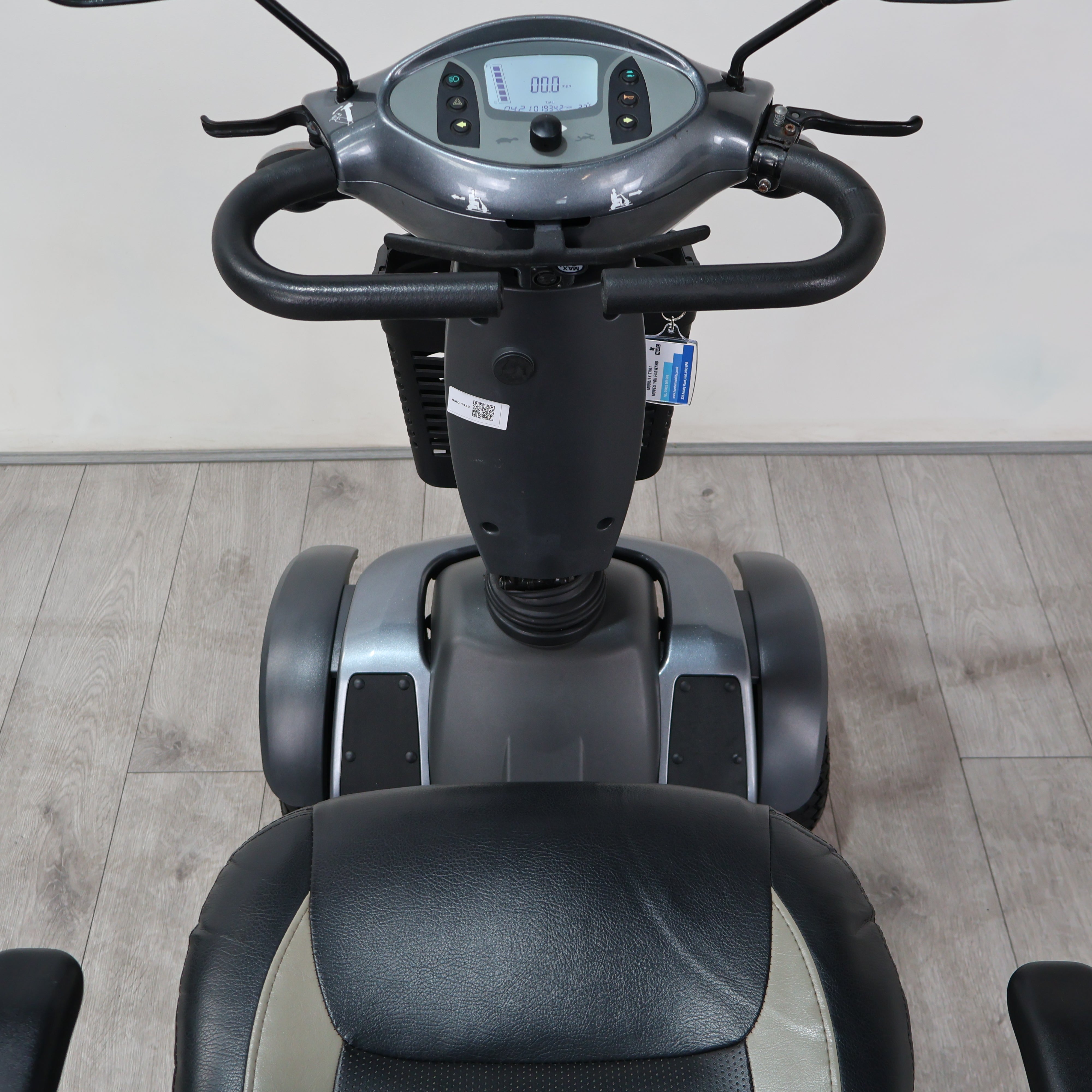 A front-facing view of a motorized three-wheel balance scooter with a digital display screen, black handlebars, and a black and gray seat, set against a light gray wall and wood-look floor.
